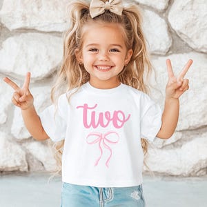 May include: A white t-shirt with a pink bow and the word "two" in pink lettering. The shirt is being worn by a young girl with blonde hair.