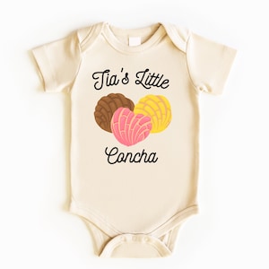 May include: A cream-colored baby onesie with the text "Tia's Little Concha" in black cursive font. Three colorful conchas are pictured above the text.