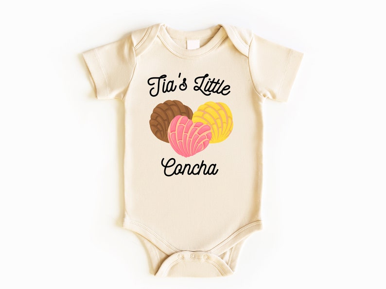 May include: A cream-colored baby onesie with the text "Tia's Little Concha" in black cursive font. Three colorful conchas are pictured above the text.