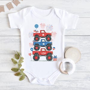 May include: White baby onesie with a patriotic design featuring three monster trucks stacked on top of each other. The trucks are red, blue, and red, with USA text and fireworks. The design includes balloons and a gift box.