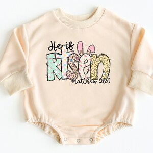 May include: A light peach colored long-sleeved baby onesie with a graphic design that says "He is Risen" with a bunny ear design and the text "Matthew 28:6".