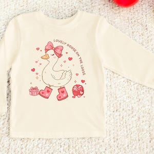 May include: Cream-colored long-sleeved shirt featuring a cartoon goose with a pink bow, boots, and hearts. The text "LOVELY GOOSE ON THE LOOSE" is printed above the goose. The design includes a gift box and a heart-shaped candy box.