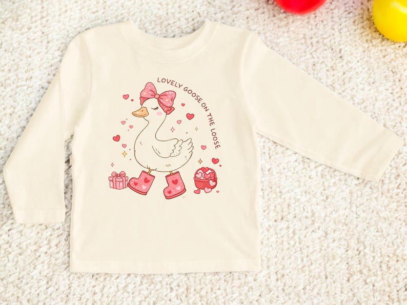 May include: Cream-colored long-sleeved shirt featuring a cartoon goose with a pink bow, boots, and hearts. The text "LOVELY GOOSE ON THE LOOSE" is printed above the goose. The design includes a gift box and a heart-shaped candy box.