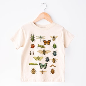 May include: Cream-colored t-shirt featuring a colorful insect illustration. The design includes various bugs like beetles, butterflies, and bees. The shirt is hanging on a wooden hanger against a white background.