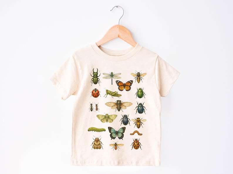 May include: Cream-colored t-shirt featuring a colorful insect illustration. The design includes various bugs like beetles, butterflies, and bees. The shirt is hanging on a wooden hanger against a white background.