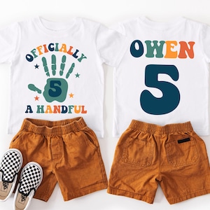 May include: White t-shirt with the text "Officially a Handful" and a green handprint with the number 5 inside. A second white t-shirt with the name "Owen" in colorful letters and the number 5 in blue. Both shirts are paired with brown shorts.