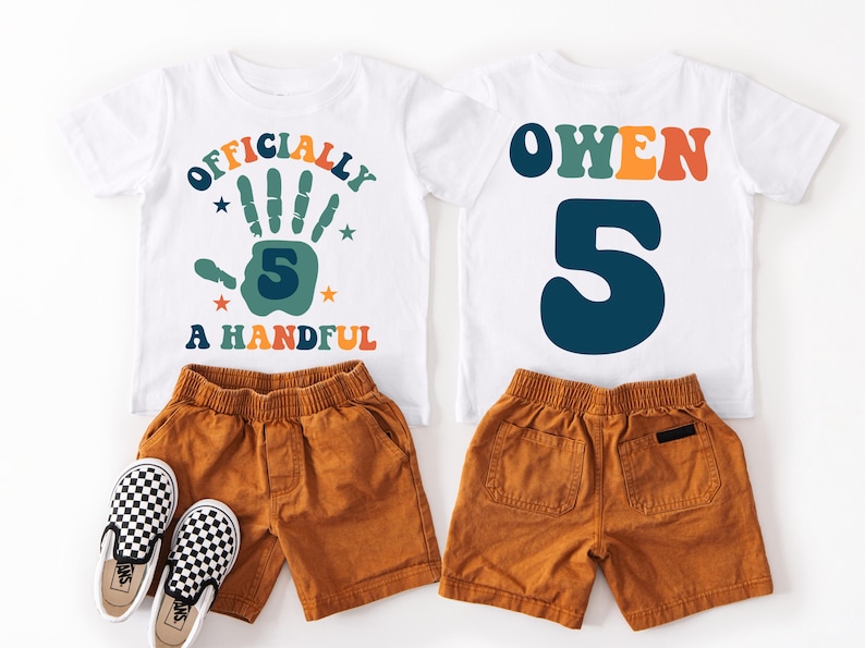 May include: White t-shirt with the text "Officially a Handful" and a green handprint with the number 5 inside. A second white t-shirt with the name "Owen" in colorful letters and the number 5 in blue. Both shirts are paired with brown shorts.