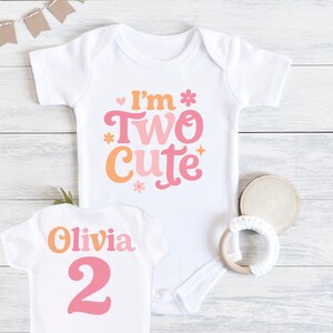 May include: White baby bodysuit with the text "I'm Two Cute" in orange and pink. A smaller white shirt with the name "Olivia" and the number "2" is also visible. A wooden teething ring and a wooden coaster are next to the shirts.