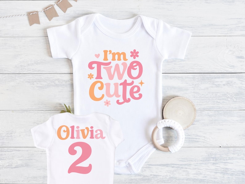 May include: White baby bodysuit with the text "I'm Two Cute" in orange and pink. A smaller white shirt with the name "Olivia" and the number "2" is also visible. A wooden teething ring and a wooden coaster are next to the shirts.