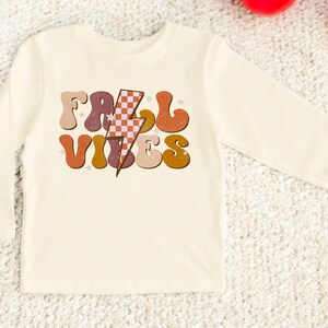 May include: A long-sleeved, cream-colored shirt with a retro-style graphic that says "Fall Vibes" in colorful, wavy letters with a checkered lightning bolt in the middle.