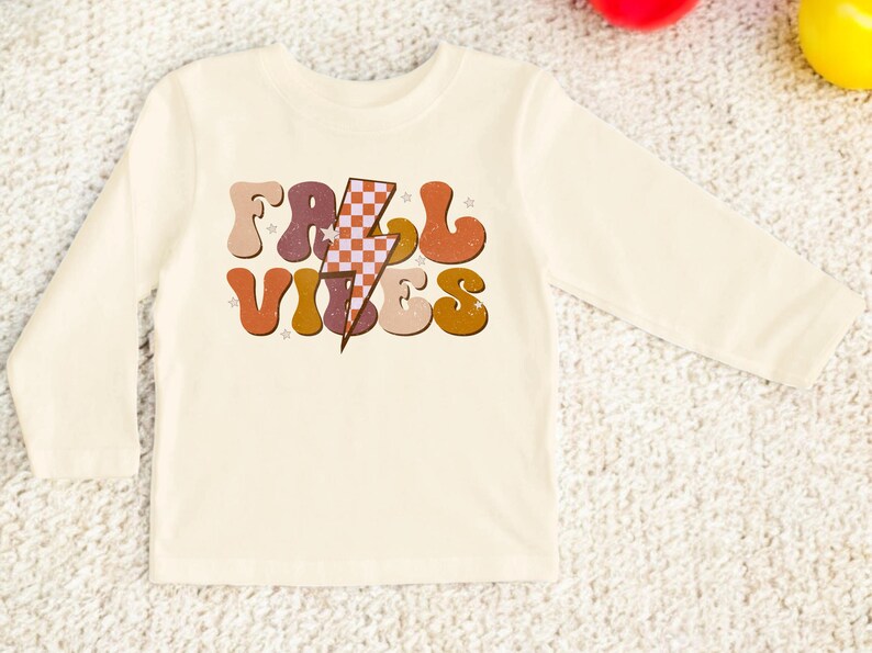 May include: A long-sleeved, cream-colored shirt with a retro-style graphic that says "Fall Vibes" in colorful, wavy letters with a checkered lightning bolt in the middle.