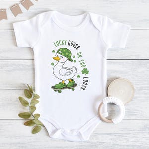 May include: White baby onesie with a cartoon goose wearing a green hat and shoes, riding a skateboard. The text "Lucky Goose on the Loose" is printed in green, with shamrock accents. A fun St. Patrick's Day themed outfit.