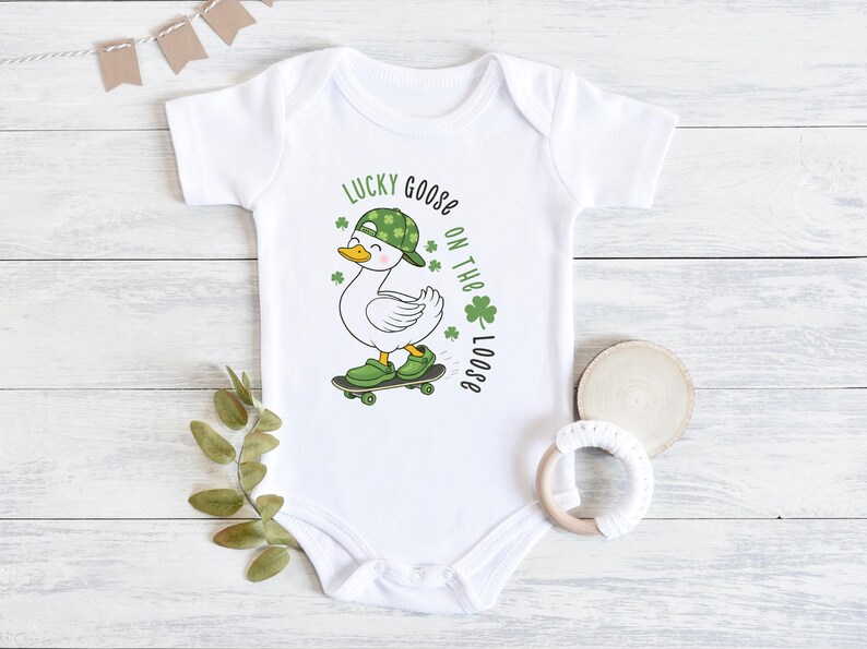 May include: White baby onesie with a cartoon goose wearing a green hat and shoes, riding a skateboard. The text "Lucky Goose on the Loose" is printed in green, with shamrock accents. A fun St. Patrick's Day themed outfit.
