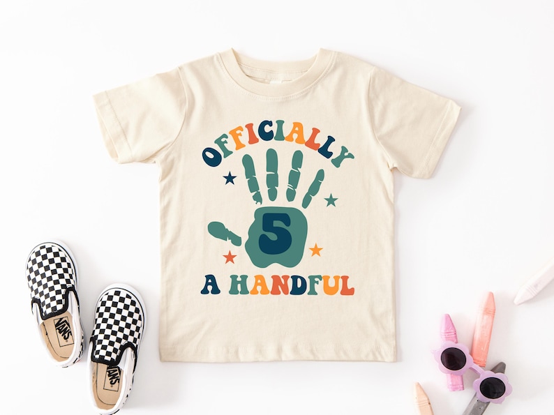 May include: A beige t-shirt with a colorful graphic that says "Officially 5 A Handful". The graphic features a handprint with the number 5 in the center.