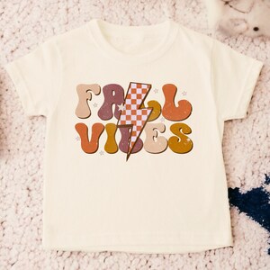 May include: A white t-shirt with a retro-style graphic that says "Fall Vibes" in colorful, outlined letters. A checkered lightning bolt is in the center of the design.