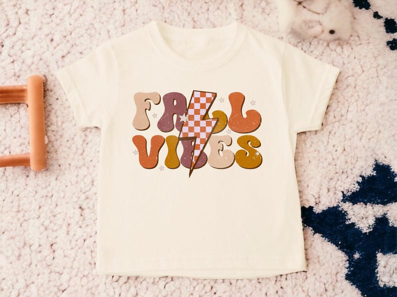 May include: A white t-shirt with a retro-style graphic that says "Fall Vibes" in colorful, outlined letters. A checkered lightning bolt is in the center of the design.