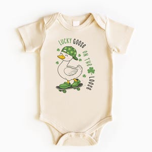 May include: Cream-colored baby bodysuit featuring a cartoon goose on a skateboard, wearing a green hat and shoes. The text "LUCKY GOOSE ON THE LOOSE" is printed above the goose, with green shamrocks. A fun St. Patrick's Day themed outfit.