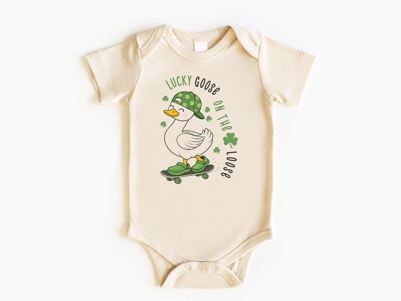 May include: Cream-colored baby bodysuit featuring a cartoon goose on a skateboard, wearing a green hat and shoes. The text "LUCKY GOOSE ON THE LOOSE" is printed above the goose, with green shamrocks. A fun St. Patrick's Day themed outfit.