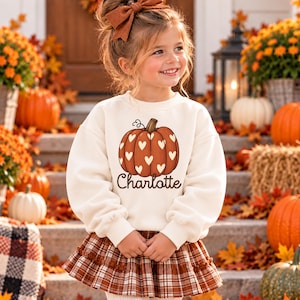 May include: A cream-colored long-sleeve sweatshirt featuring a graphic of an orange pumpkin decorated with white hearts and the name Charlotte printed in brown script lettering below the pumpkin.