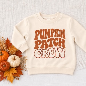 May include: A cream-colored long-sleeve sweatshirt featuring the text PUMPKIN PATCH CREW in a bold, wavy, burnt orange font centered on the chest. The garment has a ribbed crew neckline, cuffs, and hem, providing a clean and simple look.