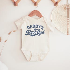 May include: Cream-colored baby onesie with the text "Daddy's Best Bud" in navy blue script. The onesie is hanging on a wooden hanger against a white background. A decorative fan and wall hooks are also visible.