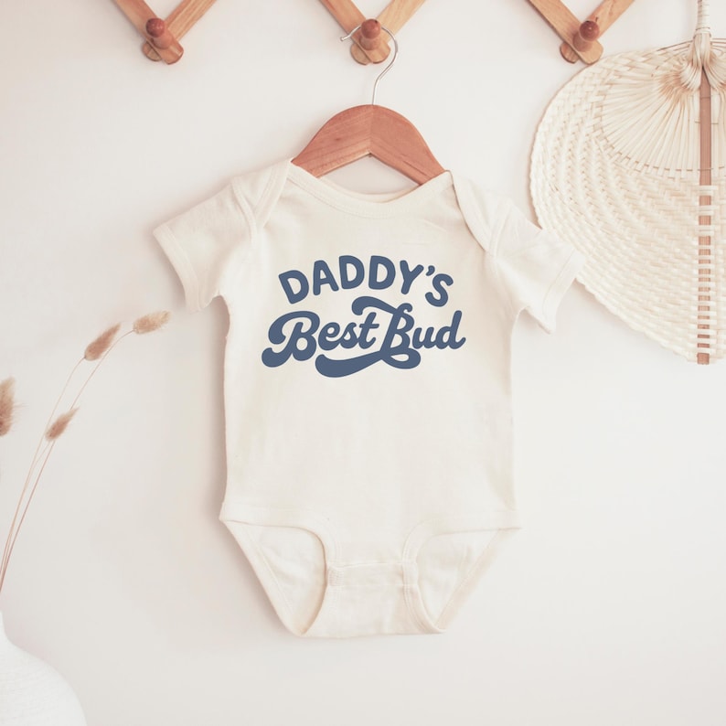 May include: Cream-colored baby onesie with the text "Daddy's Best Bud" in navy blue script. The onesie is hanging on a wooden hanger against a white background. A decorative fan and wall hooks are also visible.
