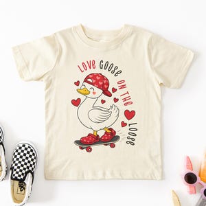 May include: Cream-colored t-shirt featuring a cartoon goose on a skateboard, wearing a red hat and shoes with heart accents. The shirt has the text "Love Goose on the Loose" in red and black. Checkered Vans slip-on shoes are on the left.