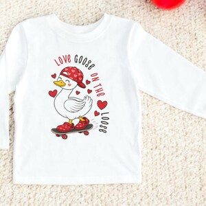 May include: White long-sleeve shirt featuring a cartoon goose on a skateboard, wearing a red hat with heart patterns and red shoes. The shirt has the text "Love Goose on the Loose" in red. A fun and playful design for kids.