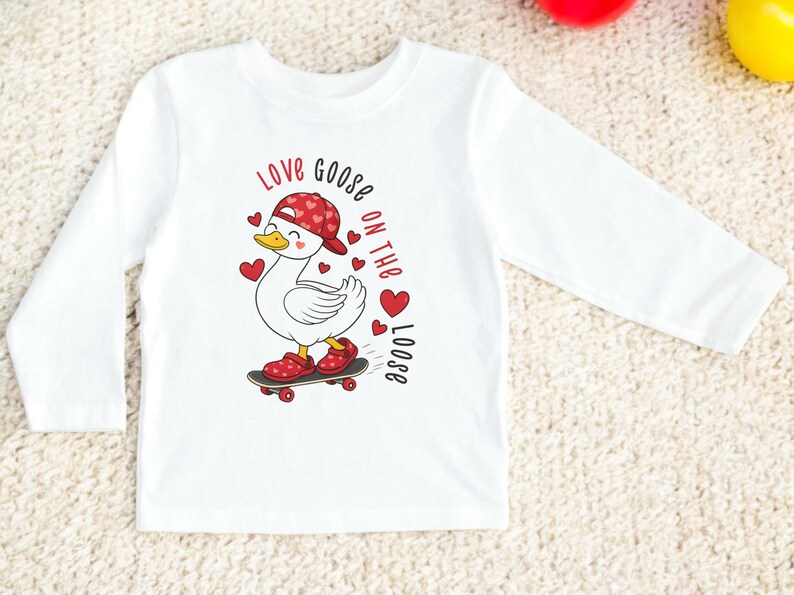 May include: White long-sleeve shirt featuring a cartoon goose on a skateboard, wearing a red hat with heart patterns and red shoes. The shirt has the text "Love Goose on the Loose" in red. A fun and playful design for kids.