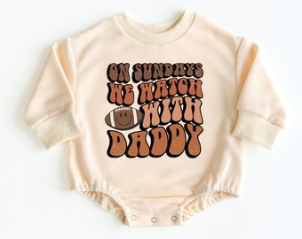 May include: A cream-colored long-sleeved baby onesie with a brown, orange, and yellow graphic that says "On Sundays we watch with Daddy" and a football with a smiley face.