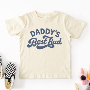 May include: Cream-colored toddler t-shirt with the text "DADDY'S Best Bud" in navy blue. The shirt is paired with black and white checkered slip-on shoes. Crayons and sunglasses are also visible.