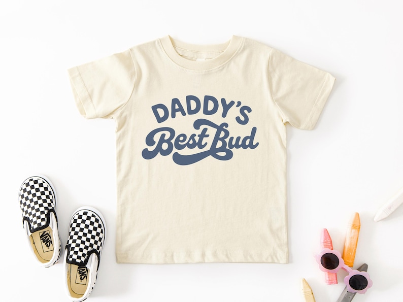 May include: Cream-colored toddler t-shirt with the text "DADDY'S Best Bud" in navy blue. The shirt is paired with black and white checkered slip-on shoes. Crayons and sunglasses are also visible.
