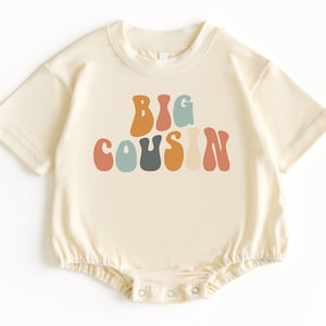 May include: A cream colored baby onesie with the words "BIG COUSIN" in colorful, retro-style lettering.