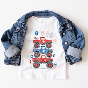 May include: A white t-shirt featuring a stack of three monster trucks in red and blue, with USA text, fireworks, and balloons. A blue denim jacket is draped over the shirt. The design is patriotic and suitable for children.