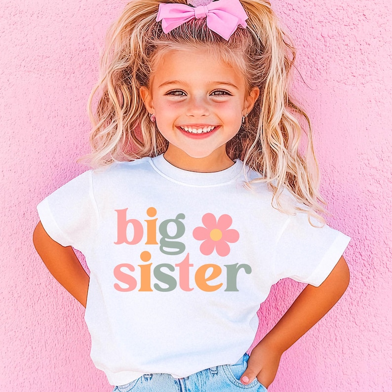 Puede incluir: Una ni&ntilde;a peque&ntilde;a lleva una camiseta blanca con las palabras "big sister" en una fuente retro colorida. La camiseta presenta un gr&aacute;fico de flores. Tiene el pelo rubio con un lazo rosa y sonr&iacute;e frente a un fondo rosa.
