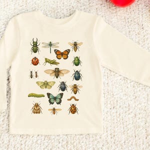 May include: Cream-colored long-sleeved shirt featuring a collection of colorful insect illustrations. The insects include a dragonfly, butterfly, ladybug, and various beetles. The shirt is made of a soft material.
