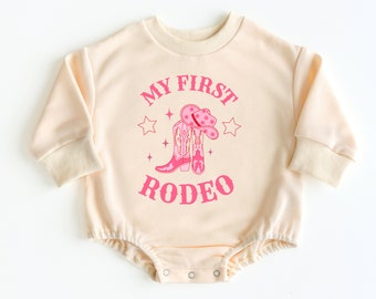 May include: A cream colored baby onesie with pink lettering that reads "My First Rodeo". The onesie has a pink graphic of cowboy boots and a hat.
