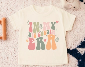 May include: A white t-shirt with a colorful graphic that says "In My Big Cousin Era" in a retro font. The graphic includes butterflies, hearts, and stars.