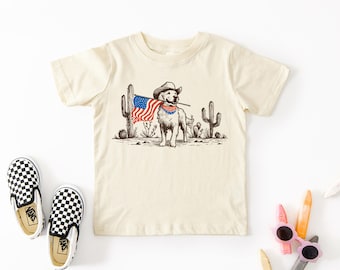 May include: A beige t-shirt with a graphic of a dog wearing a cowboy hat and bandana, holding an American flag. The dog is standing in a desert landscape with cacti.