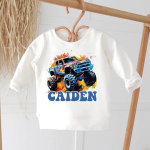 May include: White long-sleeve shirt with a colorful monster truck graphic and the name "CAIDEN" printed in blue.