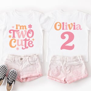 May include: Two white t-shirts with colorful text, one saying "I'm Two Cute" and the other "Olivia 2". Paired with pink shorts and black and white checkered sneakers, perfect for a second birthday celebration.