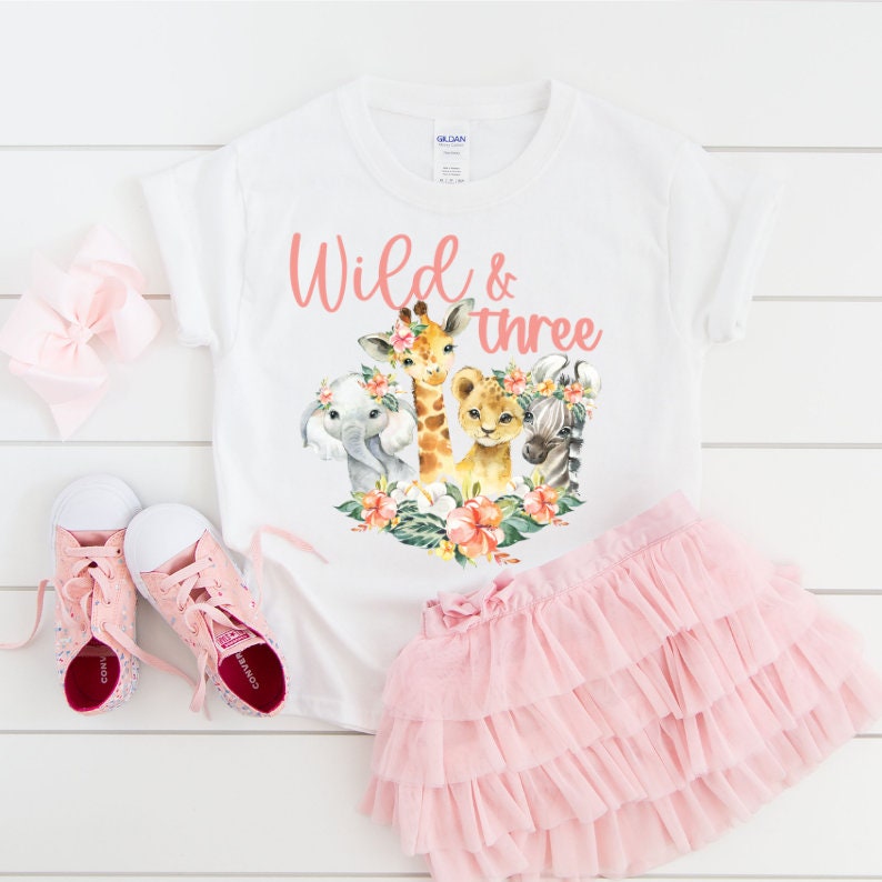 Puede incluir: Camiseta blanca con un gr&aacute;fico rosa "Wild & three" que presenta una jirafa, un elefante, un le&oacute;n y una cebra con acentos florales. La camiseta se combina con una falda de tul rosa y zapatillas rosas.