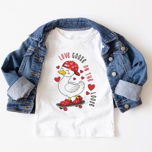May include: White t-shirt with a cartoon goose wearing a red cap and shoes, riding a skateboard. The shirt has the text "Love Goose on the Loose" and red heart accents. A blue denim jacket is draped over the shirt.