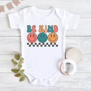 May include: White baby onesie with the text "Be Kind" in colorful retro letters. Three smiling faces with different colored eyes are below the text.