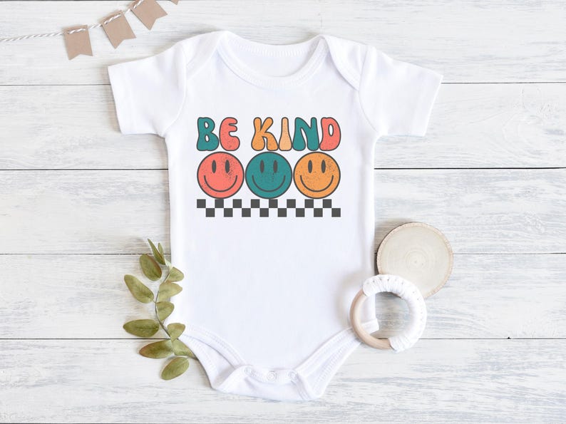 May include: White baby onesie with the text "Be Kind" in colorful retro letters. Three smiling faces with different colored eyes are below the text.