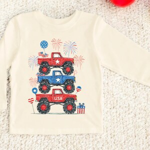 May include: Cream-colored long-sleeve shirt featuring a graphic of three monster trucks stacked on top of each other. The trucks are red and blue with black wheels, and the design includes fireworks, stars, and balloons. The bottom truck has the word "USA".