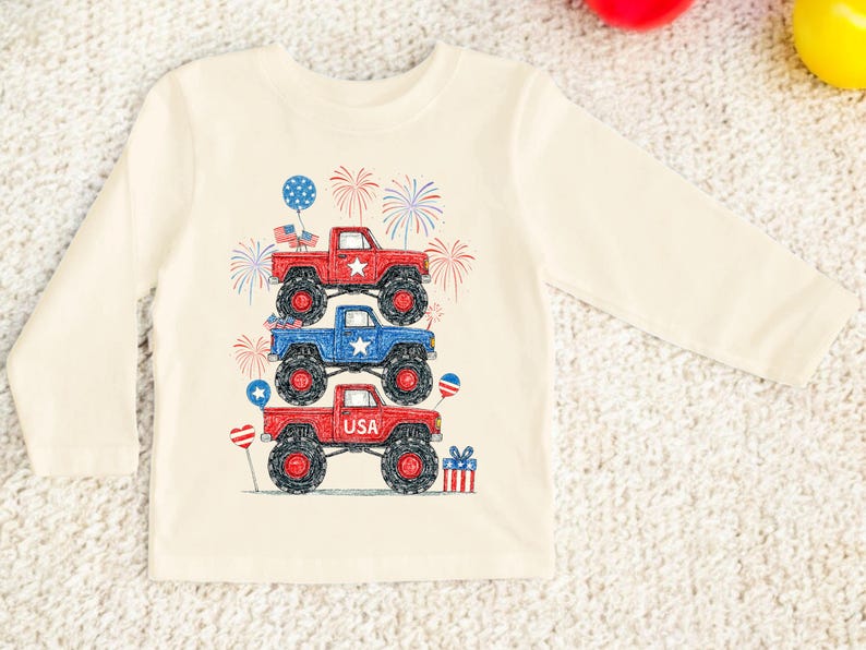 May include: Cream-colored long-sleeve shirt featuring a graphic of three monster trucks stacked on top of each other. The trucks are red and blue with black wheels, and the design includes fireworks, stars, and balloons. The bottom truck has the word "USA".
