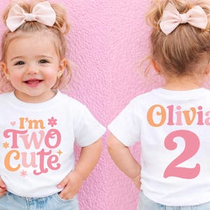 May include: A white t-shirt with the text "I'm Two Cute" in pink and orange, worn by a young child. The back of the shirt says "Olivia" and the number "2". The child is wearing a bow and jeans.