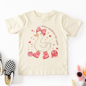 May include: Cream-colored t-shirt featuring a cartoon goose with a pink bow, heart-shaped boots, and the text "Lovely Goose on the Loose." The design includes hearts and gift boxes, perfect for Valentine's Day or a cute, whimsical look.