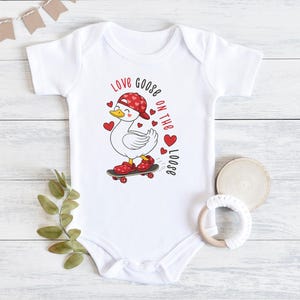 May include: White baby onesie with a cartoon goose wearing a red hat and shoes, riding a skateboard. The text "Love Goose on the Loose" is printed in red. The onesie has short sleeves and a round neckline.